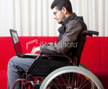 A photo of a young adult man sitting on a maroon wheelchair facing towards the left. He is wearing grayish blue jeans, a long-sleeved button down shirt with a pattern of dark gray squares and dull yellow lines, and a dark gray vest. In the background there is a red object that is possibly a couch and behind the couch there are white curtains. The man has short dark brown hair, black wire frame glasses, and a lot of light facial hair darker than a five o'clock shadow. He is using a black laptop, which is sitting on his lap